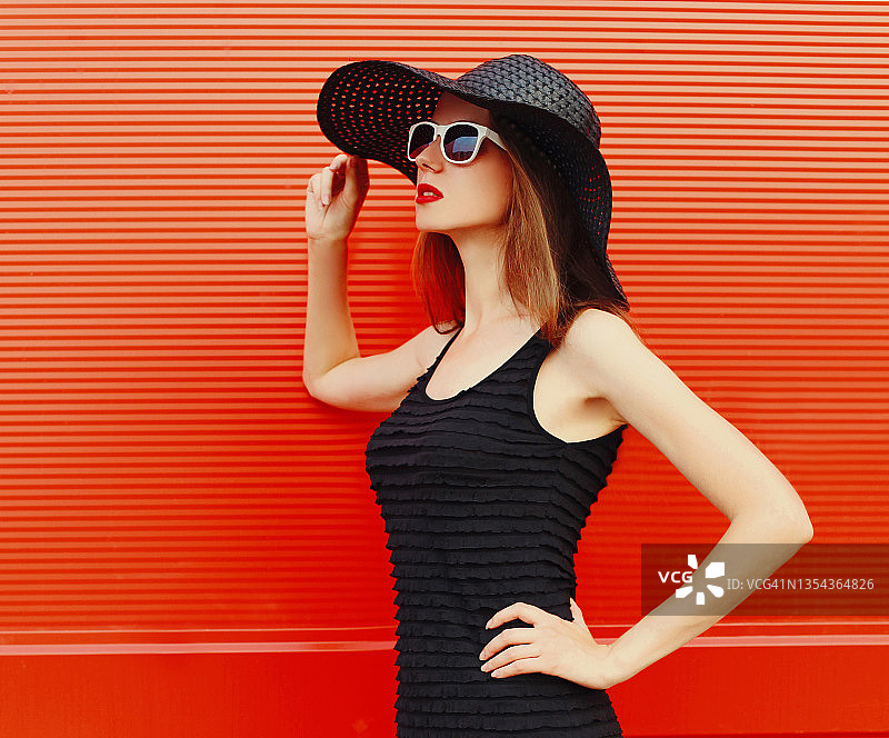 漂亮的年轻女子肖像戴着黑色夏季帽子和红色背景上的衣服图片素材