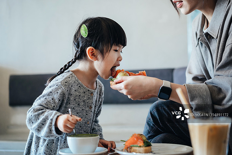 年轻的亚洲母亲和可爱的小女儿在咖啡厅吃午餐，母亲给女儿一片鳄梨吐司。家庭、饮食、生活理念图片素材