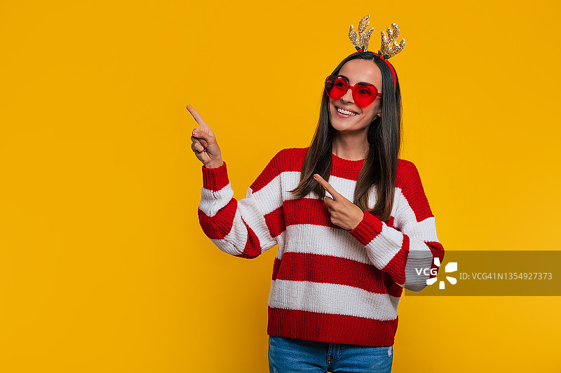 一个迷人的兴奋微笑的年轻女孩在圣诞节驯鹿鹿角和毛衣，而她指着在工作室的黄色背景图片素材