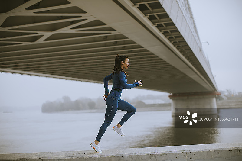 秋天的早晨，穿着蓝色运动服的年轻女子在河边跑步图片素材