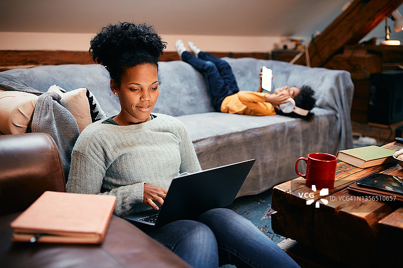 微笑的黑人母亲在家里和她的女儿在一起时使用笔记本电脑。图片素材