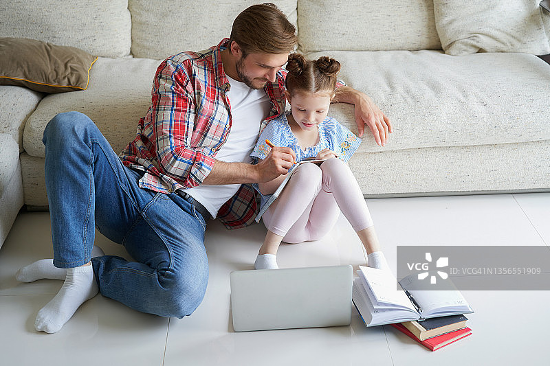 微笑的父亲和女儿坐在客厅的地板上，带着笔记本电脑，上课。图片素材