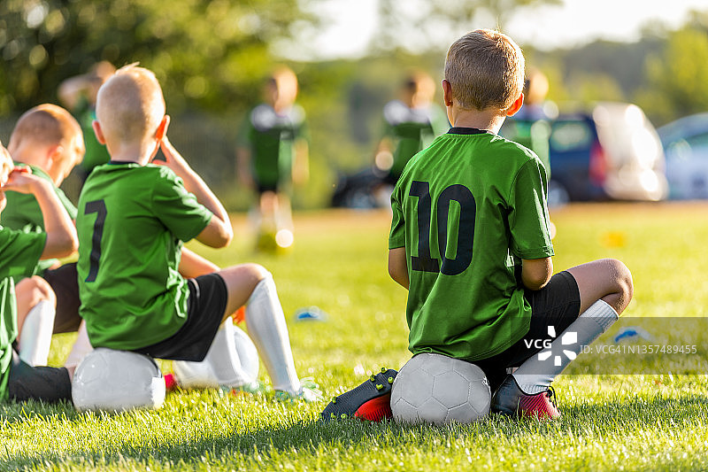 一群年轻男孩在草地上踢体育足球比赛。男孩们坐在足球上等待比赛。运动队的朋友。学校男孩在场外。穿着绿色运动衫的孩子们图片素材