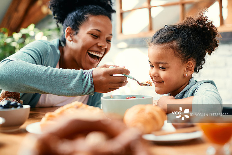 心情愉快的非洲裔美国母亲在家里吃早餐时喂她的小女儿。图片素材
