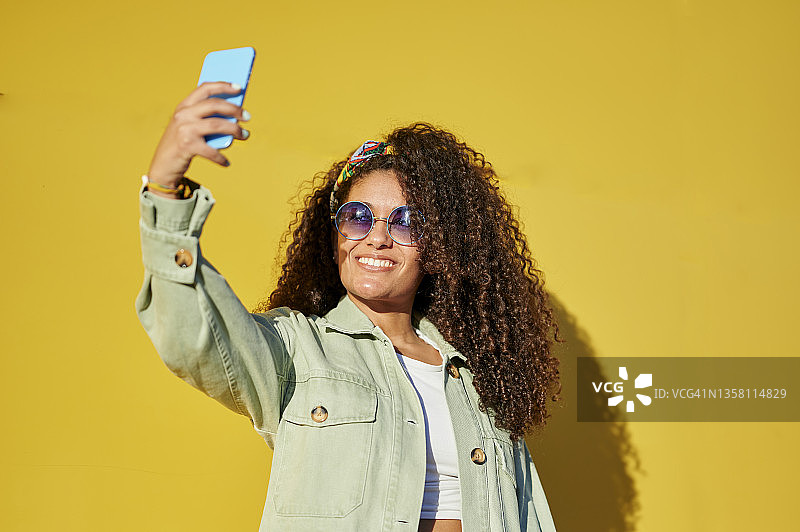 一个戴着墨镜、留着非洲式发型的女人在黄色背景下用手机自拍图片素材