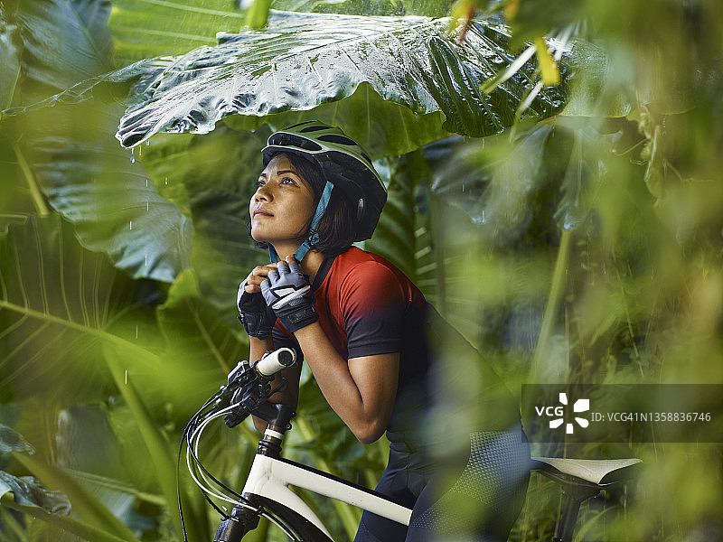 亚洲妇女坐在自行车上，调整自行车头盔，在热带植物下避雨图片素材