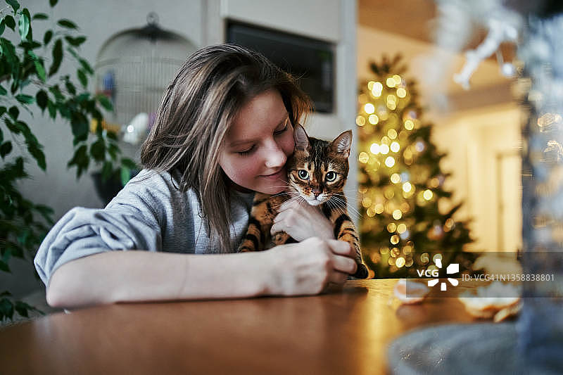 一个12岁的小女孩穿着灰色的运动衫，留着短发，正坐在家里的圆桌旁，拥抱着她心爱的条纹猫。图片素材