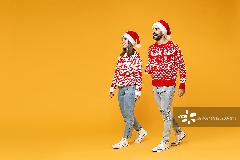 长得整整齐齐的年轻快乐的圣诞老人夫妇，朋友男人女人穿着红色毛衣，戴着圣诞帽走在黄色背景工作室肖像。欢度新年的节日理念。图片素材