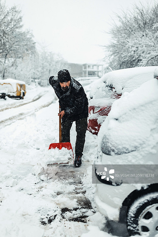 拿着雪铲的成熟男人。积雪下的汽车和街道。图片素材
