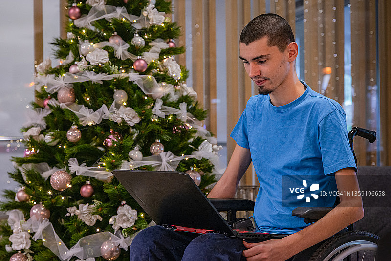 一位残疾少年——患有营养不良症，穿着蓝色t恤，坐在轮椅上，旁边是酒店大厅里一棵装饰华丽、灯火通亮的圣诞树——在电脑前工作，打电话，看着圣诞树。图片素材