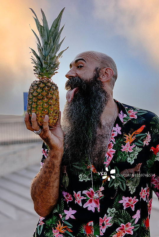一个穿着夏威夷衬衫的大胡子男子正准备舔菠萝。图片素材