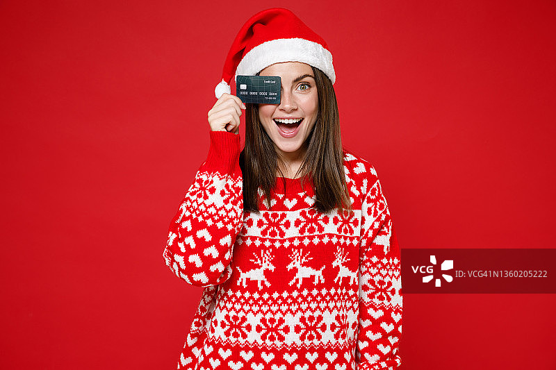 兴奋的年轻圣诞妇女穿着毛衣，圣诞帽遮住眼睛与信用卡隔离在明亮的红色背景，工作室肖像。祝新年庆佳节快乐理念。图片素材