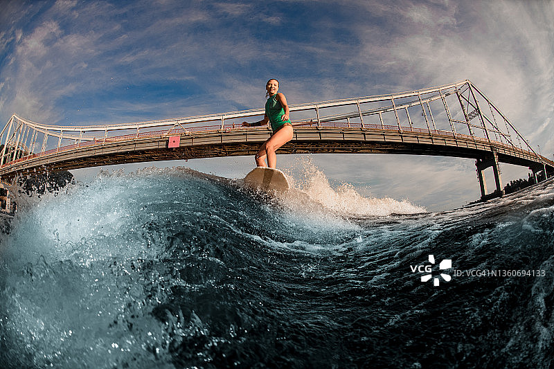 伟大的妇女wakesurfer骑在板上飞溅的波浪对桥梁。图片素材