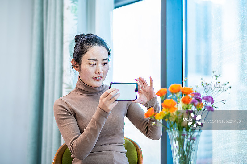 年轻的亚洲妇女在客厅用智能手机拍摄插花图片素材