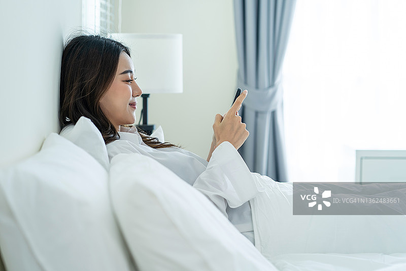亚洲美女早上在家的床上用手机聊天。快乐休闲的美女躺在卧室里享受假日周末，在屋里用智能手机和技术交流。图片素材