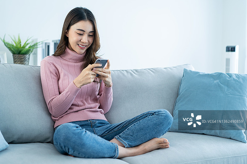 亚洲美女坐在沙发上，在家里用手机聊天。快乐迷人的年轻女孩在家里度过休闲时光，放松心情，享受在客厅用智能手机交流和讨论。图片素材