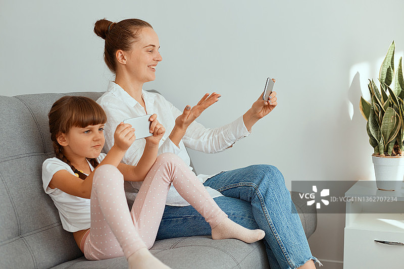 梳着发髻、穿着白色衬衫和牛仔裤的白人女性，抱着孩子坐在沙发上，用智能手机进行直播或视频通话。图片素材