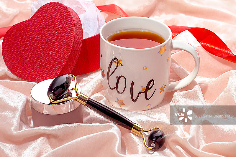 白色的茶或咖啡杯，红色的礼盒上有美容霜，黑色玉石面膜滚轮，粉色绸缎背景的缎带。图片素材