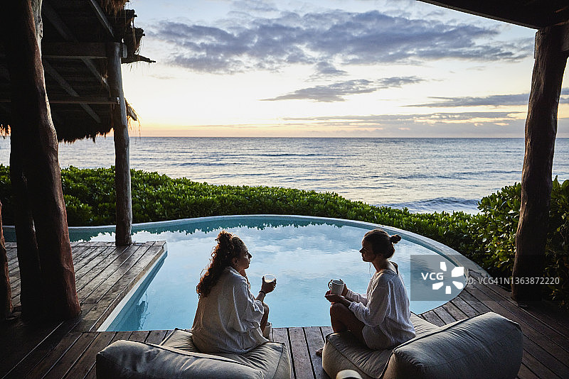 在日出时坐在热带度假胜地豪华套房的泳池边讨论的女性朋友的广泛镜头图片素材