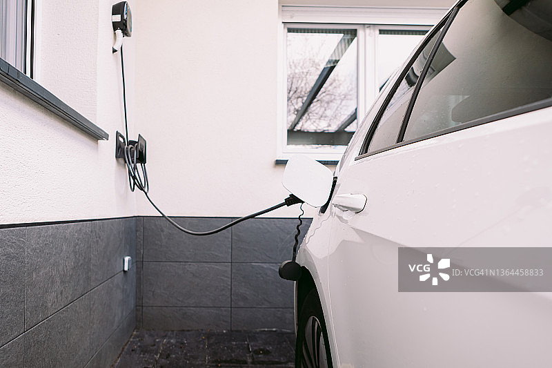 一辆电动汽车的侧视图，在一户人家外面的车库里，用电源线给电池充电。电动汽车充电、可再生能源、可持续性和交通的概念。图片素材