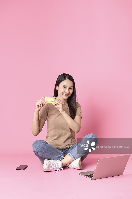 快乐女人的肖像使用笔记本电脑和智能手机与信用卡和微笑的脸，而坐在粉红色的背景图片素材
