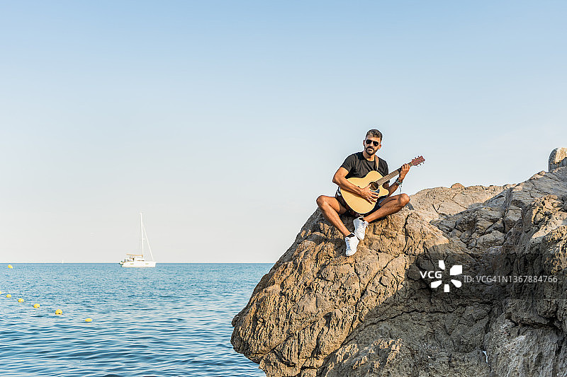 日落时分，一个年轻人坐在海滩的岩石上，悠闲地弹着原声吉他图片素材