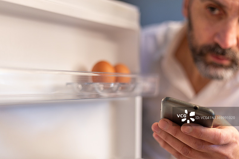 男子在冰箱里寻找食物，拿着手机。概念:点外卖图片素材