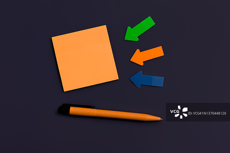 空白的橙色正方形的便利贴，钢笔和箭头的纸上深色的背景，顶视图。为未来重要事件公告留出空白图片素材