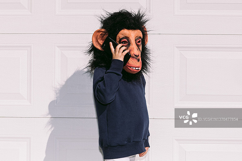 一个戴着乳胶猴子面具的孩子，在一个白色车库的门上，打电话。伪装，狂欢，奇怪，滑稽和浮华的概念。图片素材