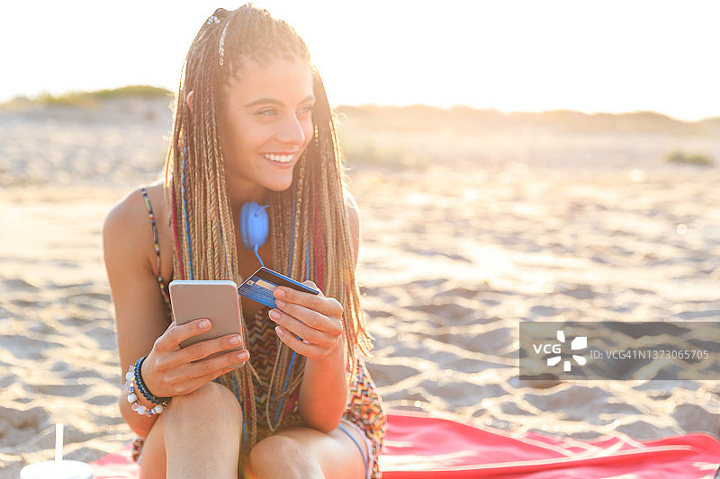 美丽微笑的女孩扎着辫子坐在沙滩上与她的手机在日落，持有信用卡和网上支付，网上购物和数字购买，在她的假期图片素材