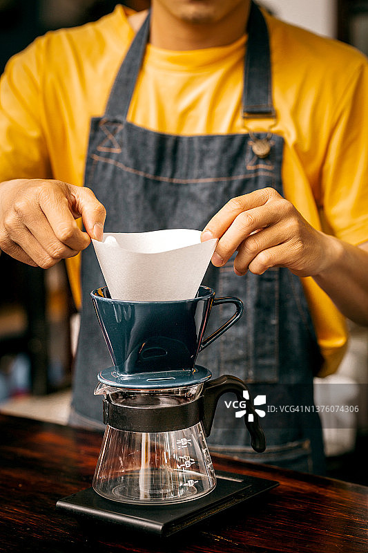 年轻的亚洲男咖啡师，穿着黄色衬衫和蓝色围裙，用卡丽塔咖啡机煮咖啡图片素材
