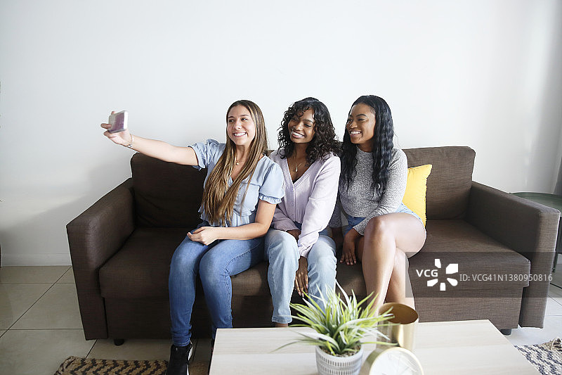 一群多种族的年轻女性，穿着休闲的衣服，在新公寓的沙发上愉快地自拍。图片素材
