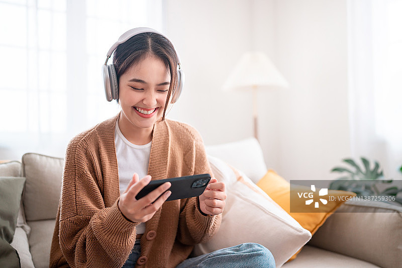 快乐的年轻亚洲女性放松和听音乐和看电影的手机与耳机，而坐在沙发上在家里早上图片素材