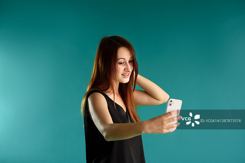 快乐的红发年轻女子享受移动通信和现代技术的肖像在蓝色背景上摆姿势。免费空间为您的广告内容。图片素材