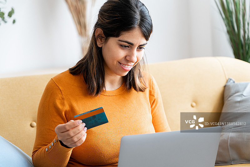 漂亮的黑发女人坐在沙发上开心地拿着她的新信用卡，准备在网上购物。图片素材
