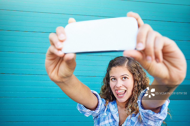 美丽的卷发年轻女子与她的手机相机在一个蓝绿色的背景与复制空间自拍图片素材