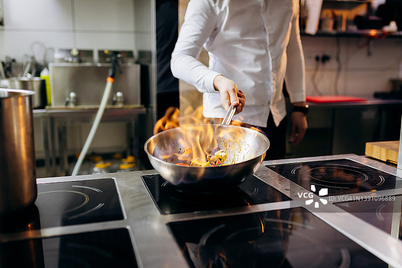 海夫在专业厨房里拿着煎锅和火。厨师的手拿着铁锅，在炊具上准备食物。图片素材