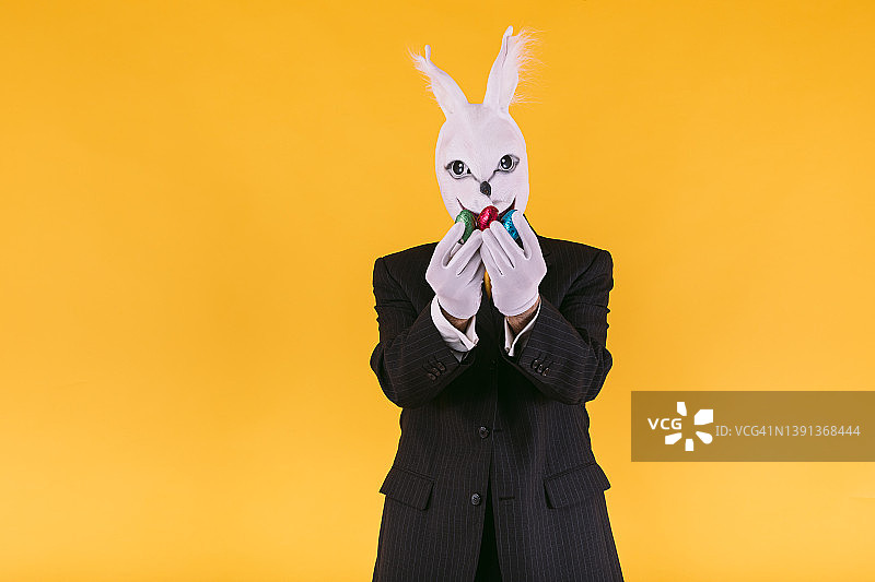 伪装的人戴着兔子面具，穿着西装外套，背心和领带，拿着复活节彩蛋，在黄色的背景上。嘉年华，派对，复活节和庆祝的概念。图片素材