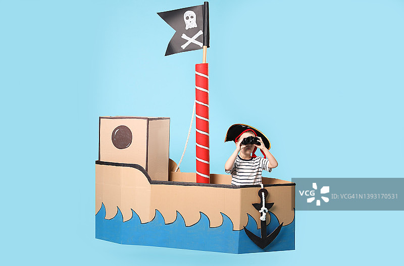 小男孩玩双筒望远镜在海盗纸板船在绿松石背景图片素材