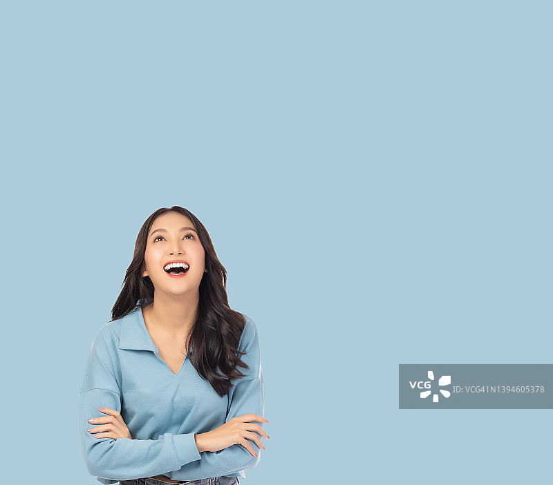 快乐的亚洲女孩抱着双臂，看着上面站在淡蓝色背景和复制空间自信的女性得到快乐和放松图片素材