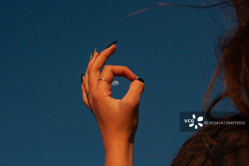 一个女人的手用长长的指甲在湛蓝清澈的天空上做着完美的手势&用手指做的戒指上有月亮图片素材