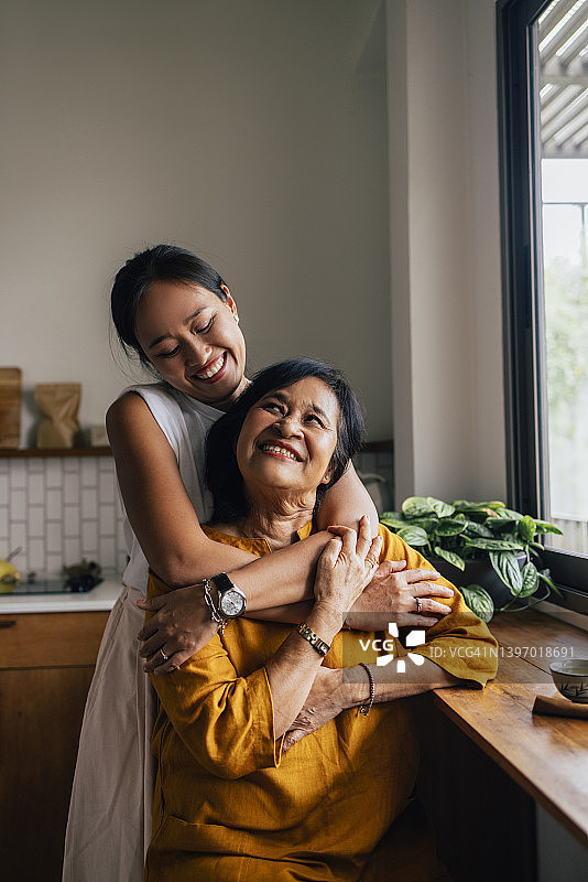 亚洲两家人的肖像，母亲和女儿，坐在厨房，女儿拥抱她的母亲充满爱和奉献。图片素材