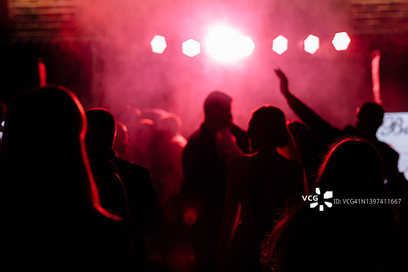 在明亮的红色舞台灯光前欢呼的人群。人们在迪斯科夜总会或音乐节的音乐会上跳舞的剪影。图片素材