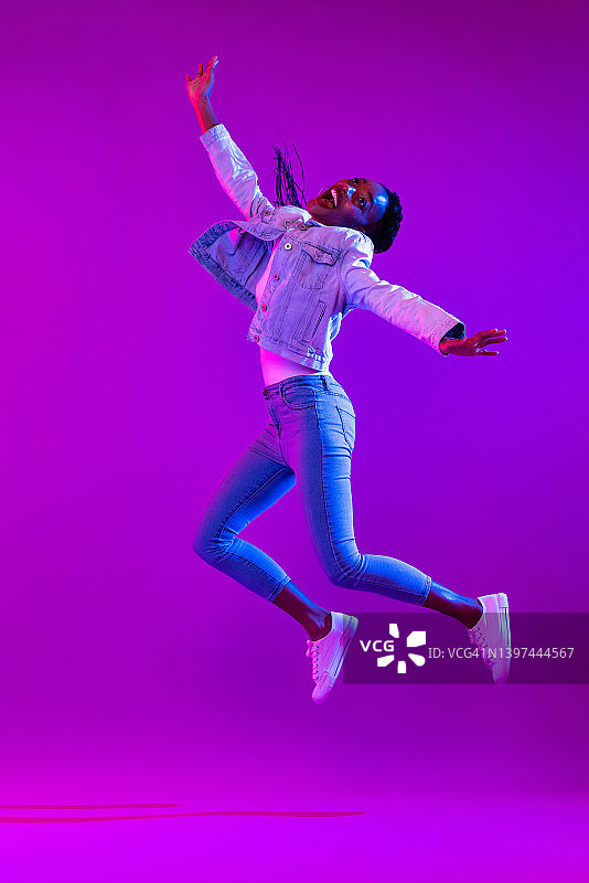 欣喜若狂的非裔美国女人在现代霓虹灯工作室背景下跳跃的全身肖像图片素材