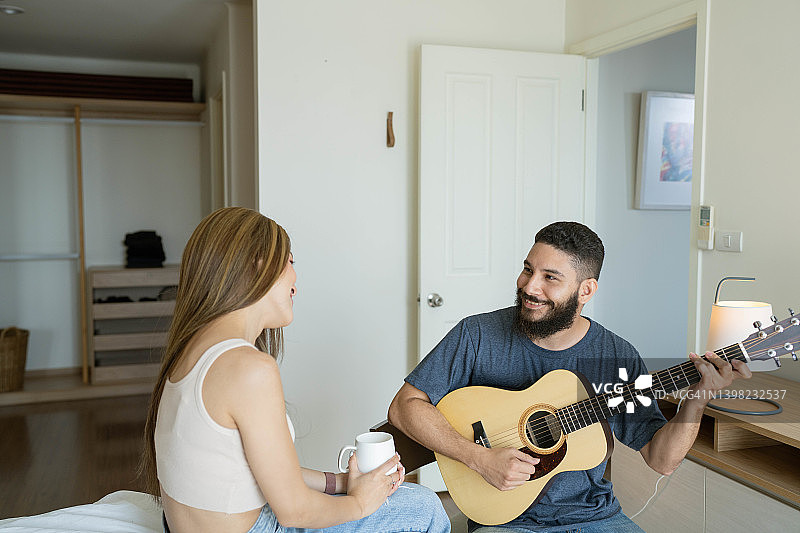 幸福的年轻的多民族夫妇喜欢唱歌和吉他，而坐在卧室在早上。图片素材