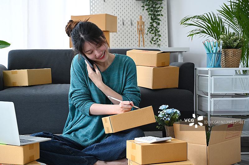 微笑的亚洲女人网上卖家收到订单在手机上，并准备产品的包裹盒交付。图片素材