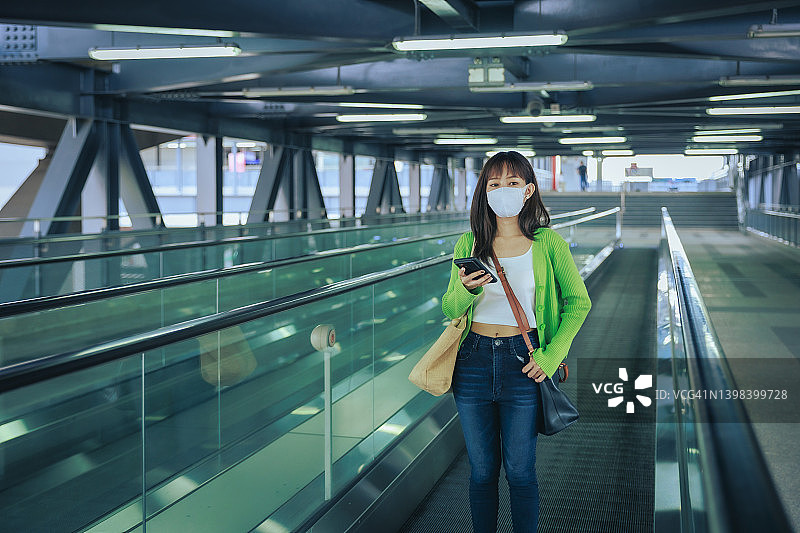 年轻的亚洲女商人戴着口罩，在机场的自动扶梯上用手机阅读电子邮件。商务旅行的概念。图片素材