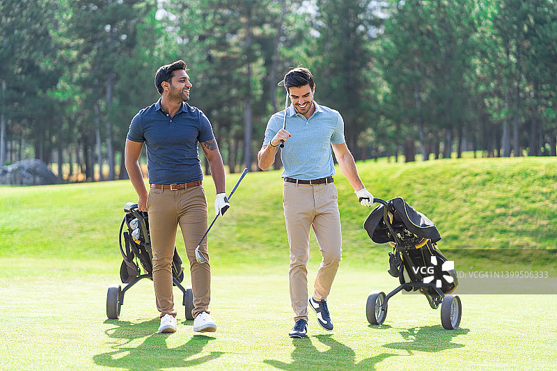 在高尔夫球场上，年轻的高尔夫球手们拿着高尔夫球杆，拉着带轮子的高尔夫球袋，一边说话一边微笑。图片素材