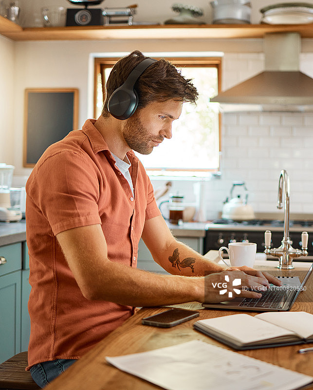 年轻认真的白人商人戴着耳机，一边听音乐，一边独自在家使用笔记本电脑。一名男性商人在家里的厨房工作时，正在笔记本电脑上打字图片素材