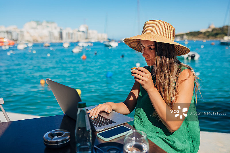 商务女性一边喝咖啡一边远程工作，坐在离海不远的咖啡店里图片素材